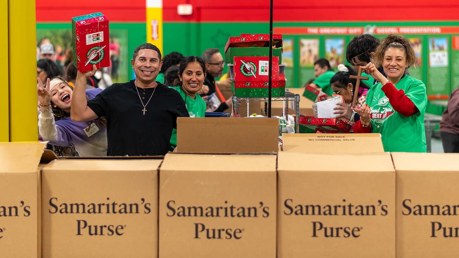 Group of volunteers smiling and holding up shoeboxes and a wooden cross standing at a processing station