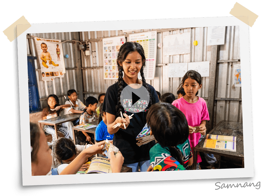 Samnang smiling and handing out The Greatest Journey books and pencils to children sitting at desks