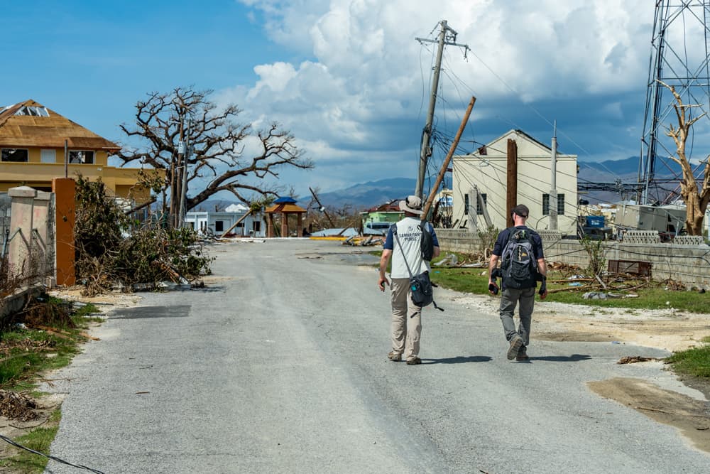 Two Samaritan’s Purse workers walk down a damaged street lined with debris and broken power poles in Jamaica.