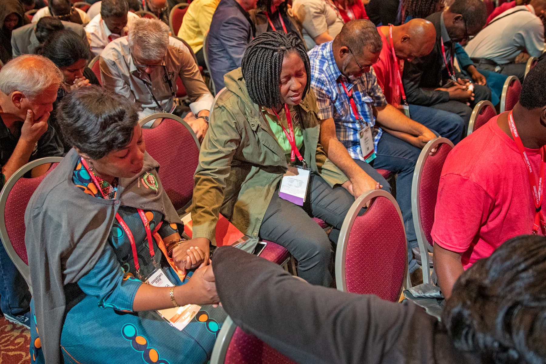 Group of people sitting in chairs with heads bowed in prayer while holding hands