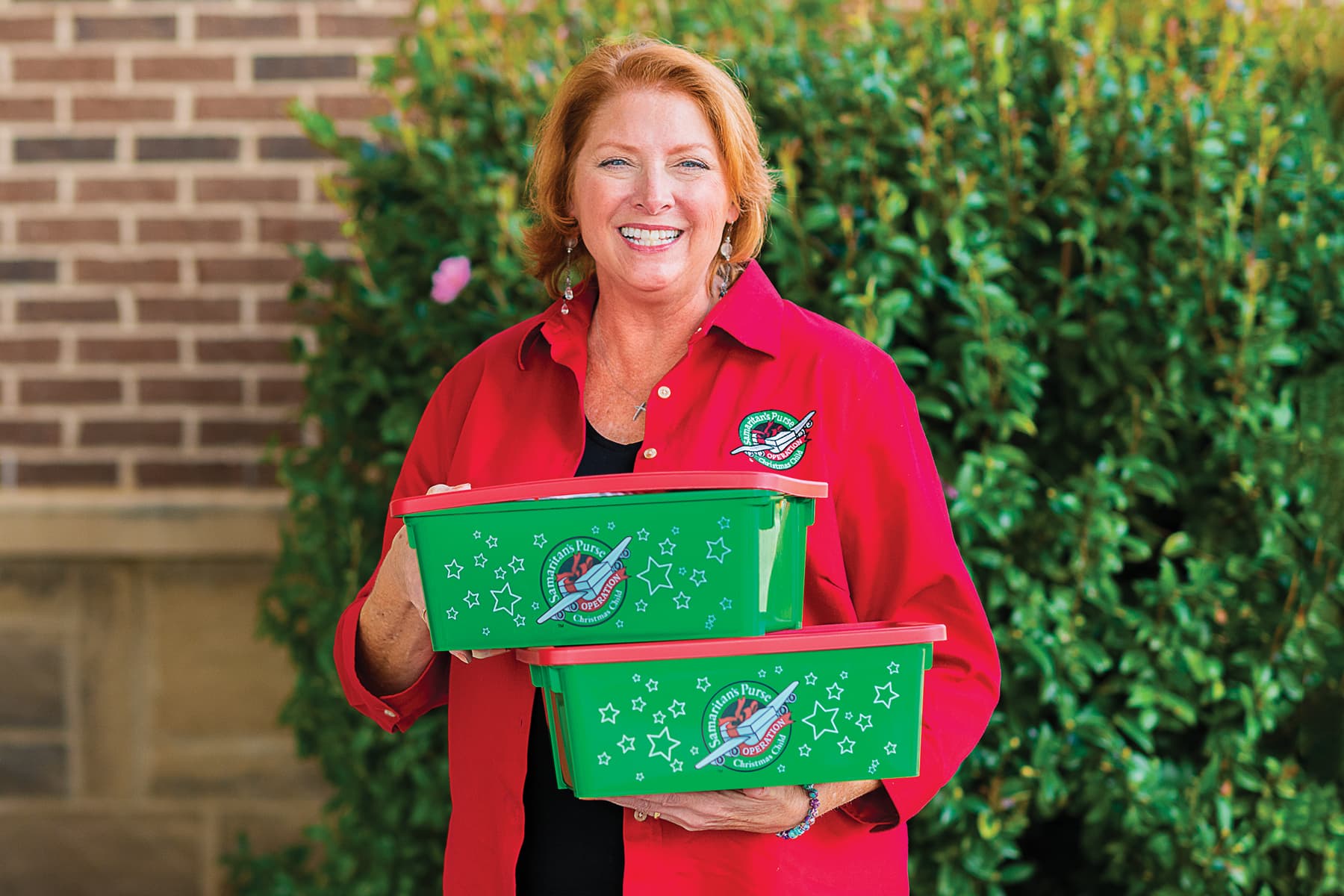 Female volunteer in a red Operation Christmas Child shirt standing outside smiling and holding two shoeboxes