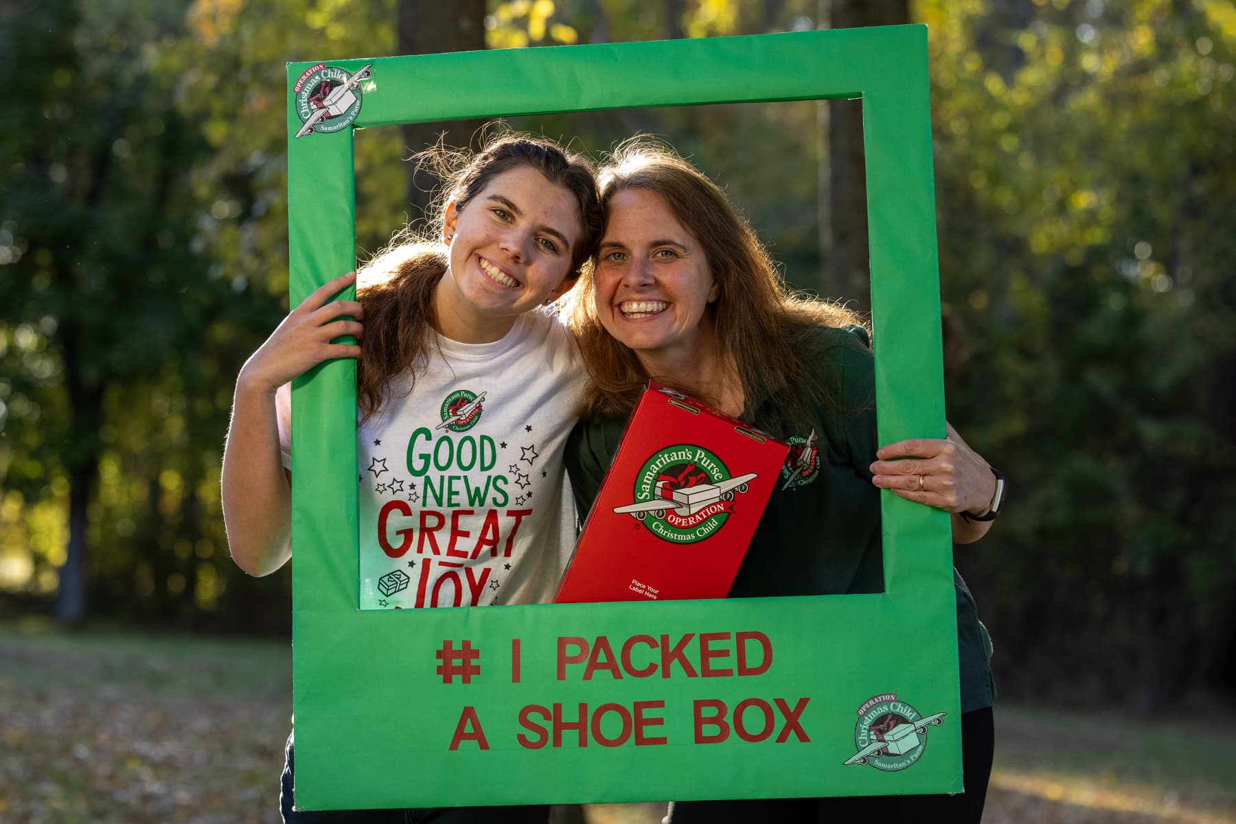 Two ladies posing for a photo outside while holding up a square, green prop with text on the bottom that reads: #I PACKED A SHOE BOX