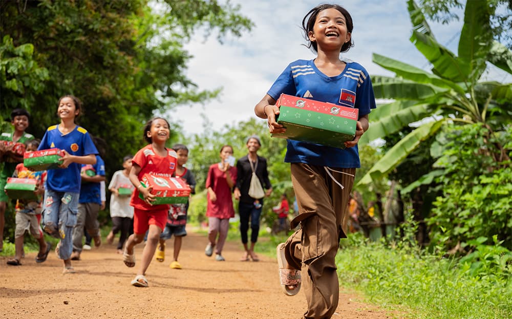 Kids smiling and running down a dirt road with Operation Christmas Child shoeboxes in hand