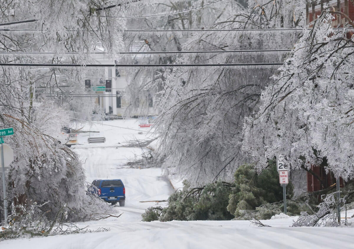 A blue truck navigates down an ice and snow covered street with trees broken and hanging down on both sides of the road.