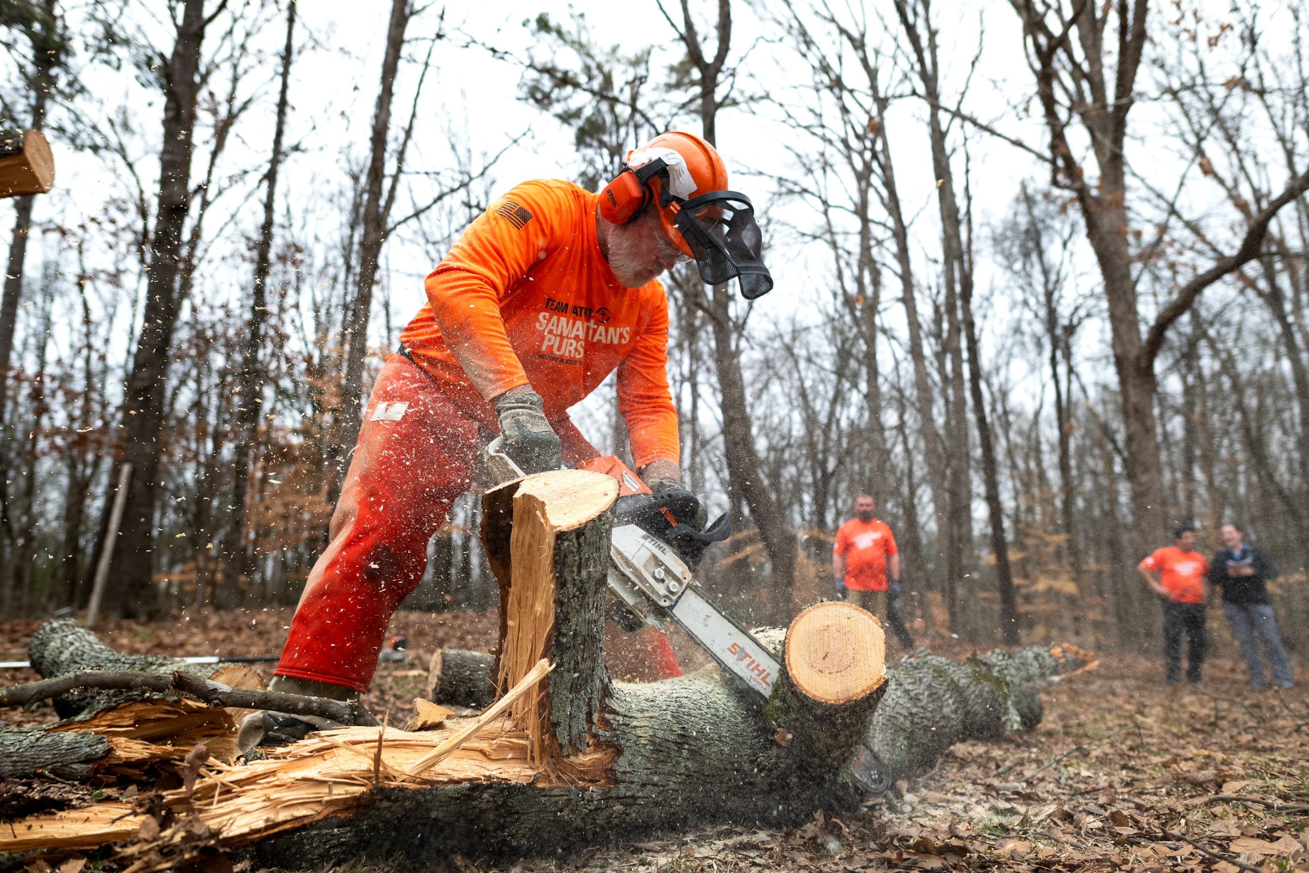 A man wearing an orange Samaritan's Purse shirt standing over a fallen tree dressed in orange protective tree cutting gear cutting into a limb with a chainsaw